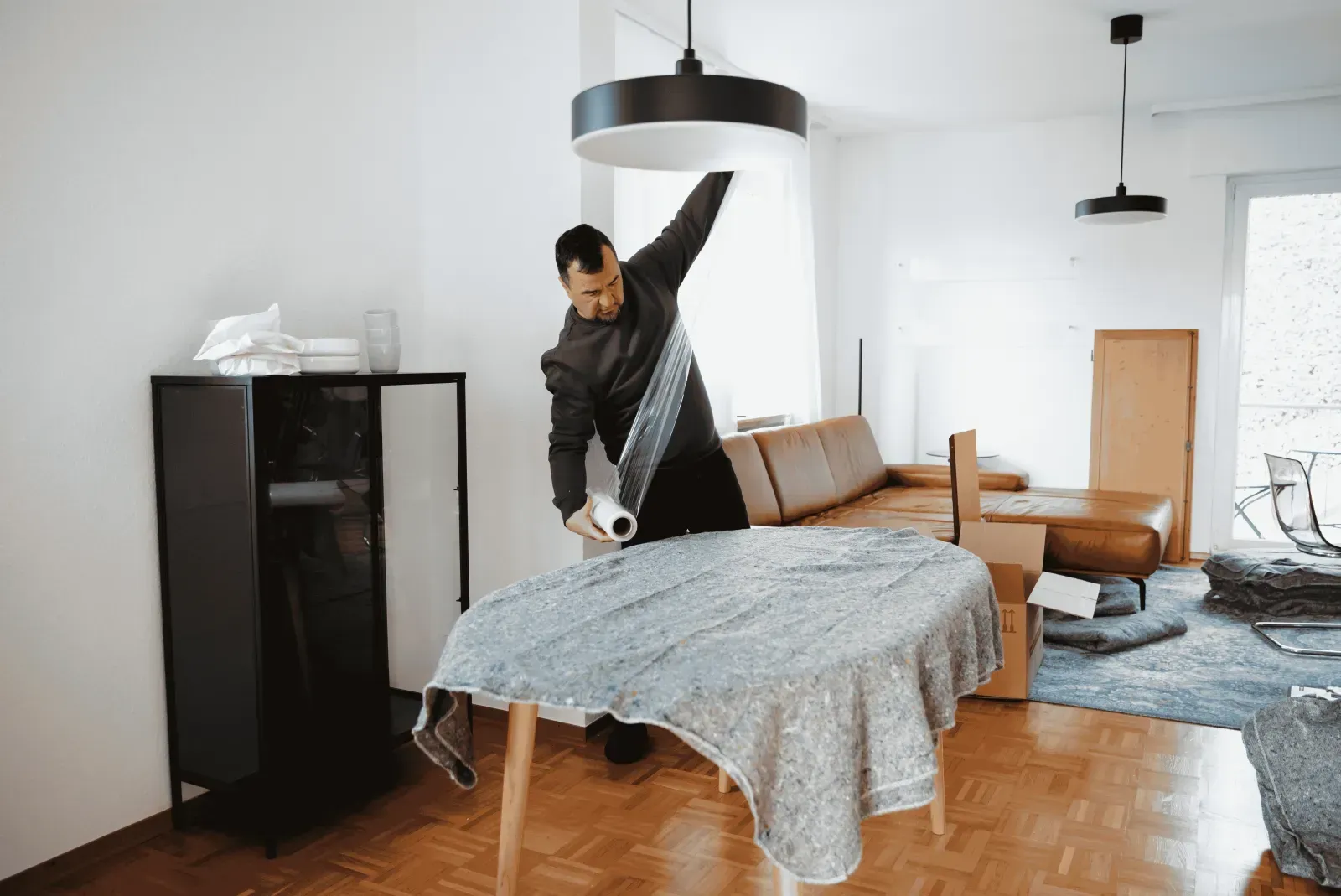 Ein Mann wickelt Stretchfolie um einen mit einer Umzugsdecke geschützten runden Tisch.