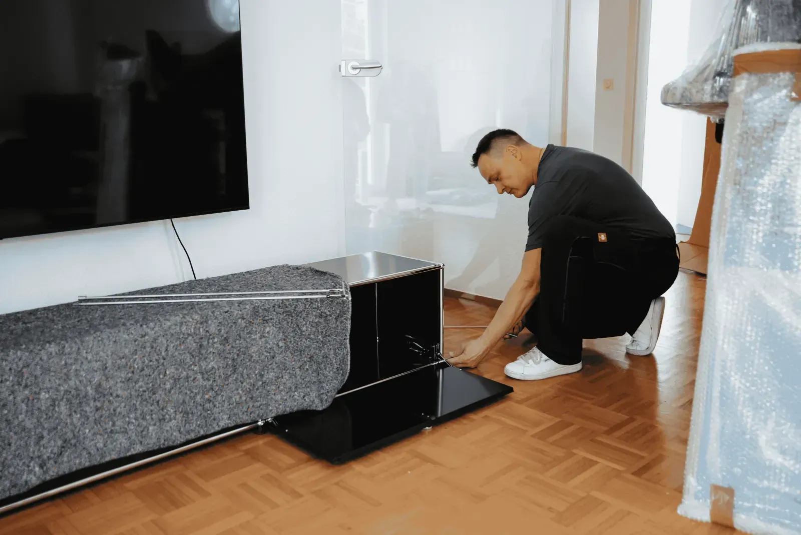 Ein Mann kniet neben einem schwarzen Möbelstück, das teilweise mit einer Umzugsdecke geschützt ist.