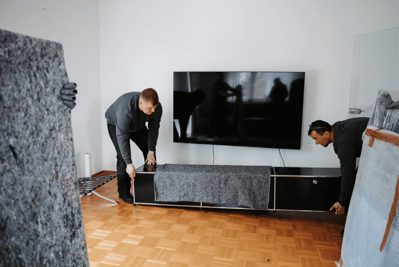 Zwei Umzugshelfer tragen ein teilweise mit einer Umzugsdecke geschütztes Sideboard in einer Wohnung.